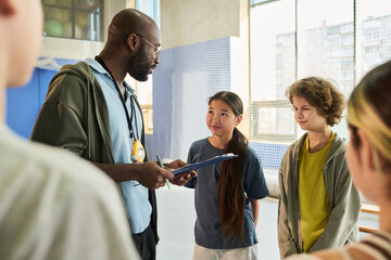 Black man coach holding clipboard talking to diverse group of teenagers including Asian girl and Caucasian boy in sports facility during team meeting, other teens listening