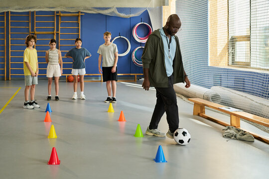 Black man demonstrating soccer dribbling skills to group of four children standing in line, watching attentively in gymnasium with colorful cones arranged on floor