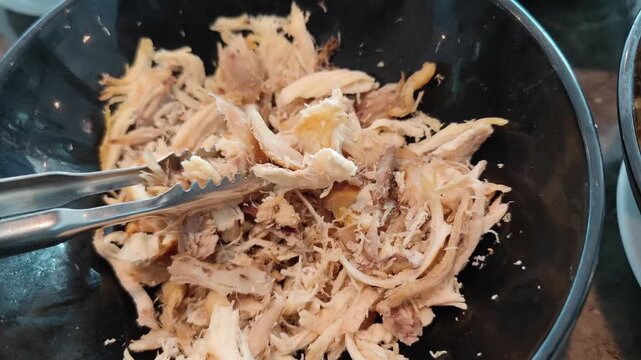 Close-up view of freshly shredded cooked chicken in a black container, held with metal tongs. Detailed texture of tender chicken meat, perfect for concepts related to food preparation, cooking process