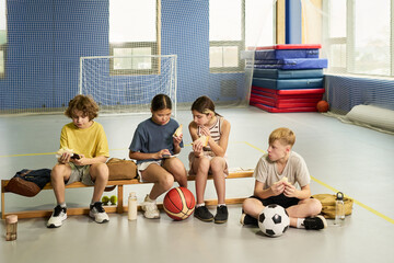 Group of four children sitting on bench in gymnasium eating snacks after sports activity, two girls and two boys, one Caucasian boy holding soccer ball, all appearing relaxed and focused