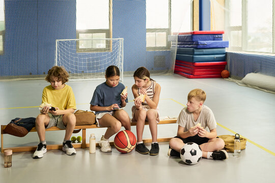 Group of four children sitting on bench in gymnasium eating snacks after sports activity, two girls and two boys, one Caucasian boy holding soccer ball, all appearing relaxed and focused