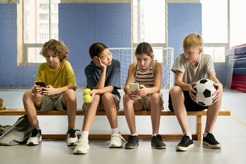 Group of four teenagers sitting on bench using smartphones and holding sports equipment, including tennis balls and soccer ball, multiethnic boys and girls interacting indoors
