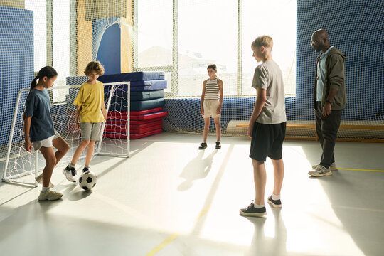 Group of multiethnic children and teenagers playing soccer indoors while Black man coach supervising, children focusing on ball, sunlight streaming through large windows in background