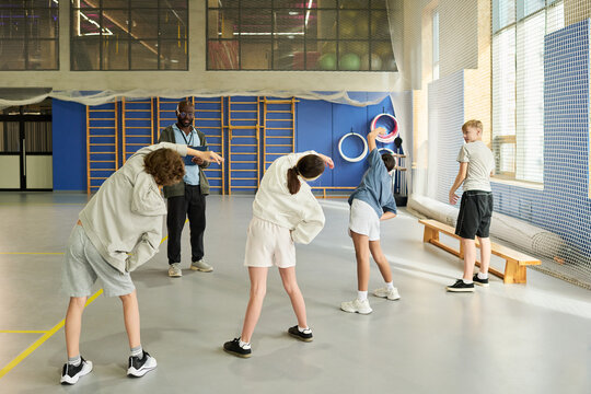 Group of multiethnic teenagers exercising under supervision of Black man coach in gym, teenagers performing side stretching together, coach observing and instructing physical activity