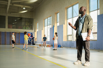 Black man coaching group of multiethnic children practicing basketball drills in gymnasium, holding clipboard and whistle, children standing with basketballs listening attentively