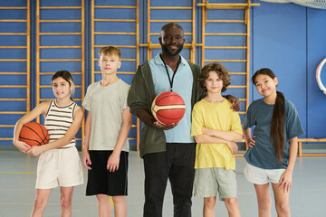 Black man standing with four teenagers in gym holding basketballs, group smiling and posing for camera, two Caucasian boys and two girls of different ethnicities standing together