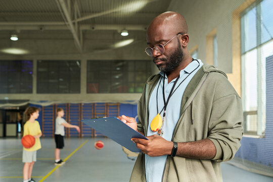 Black man holding clipboard and writing while standing in gymnasium, two Caucasian boys in background dribbling basketballs, coach supervising youth basketball practice