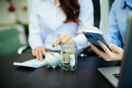 Two women managing savings with coins, jars, piggy bank and calculator at office desk. Finance, budgeting, money planning and smart financial