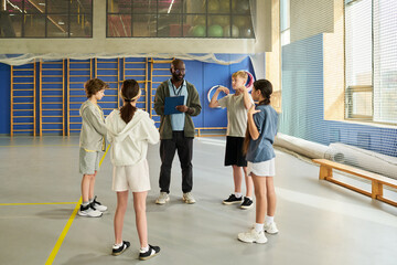 Black man instructing group of four children in sports gym, children standing in semicircle listening attentively, sports equipment and gymnasium background visible