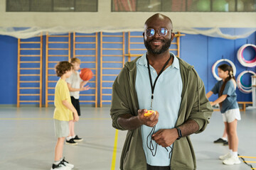 Portrait of Black man holding stopwatch and smiling at camera in gymnasium, while group of multiethnic children playing basketball in background during physical education class
