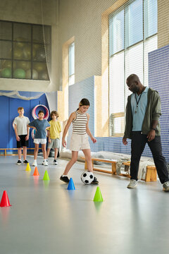 Caucasian girl dribbling soccer ball through cones while Black man supervising, group of children waiting in line behind, indoor gym setting, physical education activity
