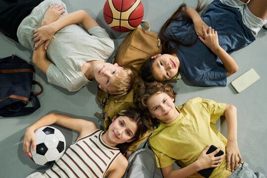 Group of four teenagers, two Caucasian boys, one Caucasian girl, one Asian girl, lying on floor in circle, smiling and looking up, surrounded by soccer ball, basketball, backpacks, smartphone
