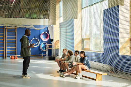 Black man standing and holding tablet while instructing group of four teenagers sitting on bench in gymnasium, sports equipment and netting visible in background