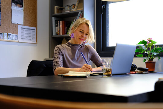 Woman working from home writing in notebook and using laptop - Powered by Adobe