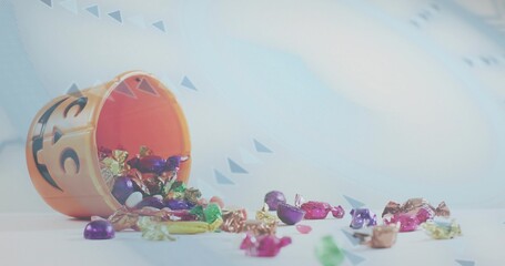 Tipping orange jack-o'-lantern bucket spilling foil candies across pale tabletop, confetti backdrop