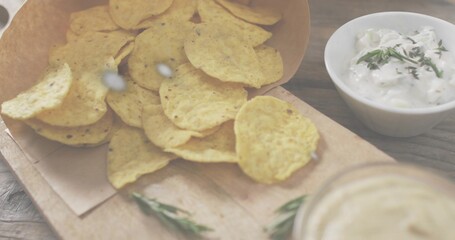 Spilling corn tortilla chips from paper cone onto wooden board on rustic tabletop, with herb dip