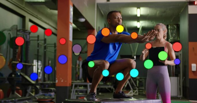Performing box squat, trainee in blue-shirt using plyo-box in weight room, trainer holding tablet