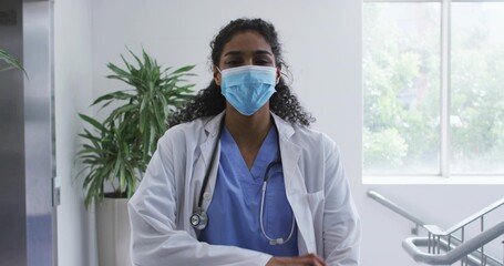 Standing doctor folding arms facing camera in clinic hall, blue scrubs, lab coat, mask, stethoscope