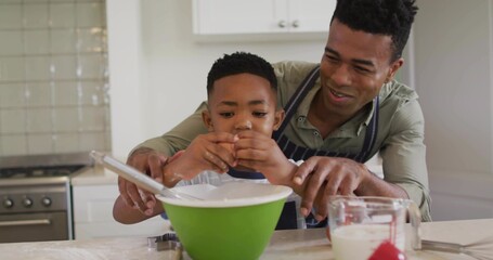 Guiding father and son cracking egg over green mixing bowl in kitchen, with whisk and aprons