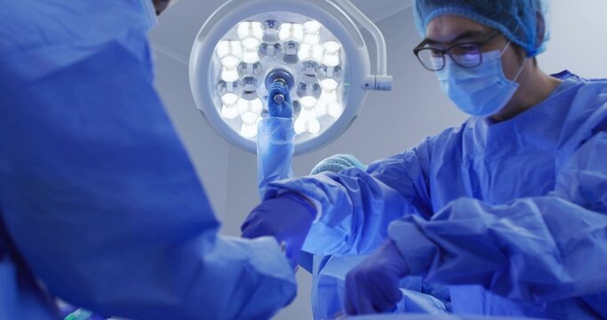 Operating surgical team adjusting instruments under LED surgical light in OR, wearing gowns, masks