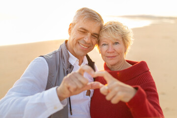 Happy senior couple creating heart hands gesture on beach