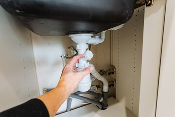 hand adjusting sink trap under cabinet, closeup of fingers and connectors while tightening joint to stop drip, practical diy plumbing task with tools and focused maintenance action