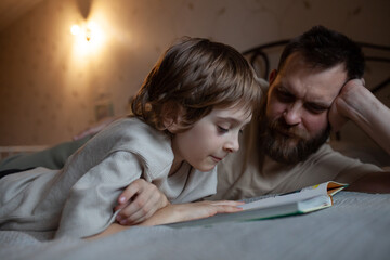 Attentive boy reading book with dad lying on bed. Kids read with interest and pleasure concept. Engaging children to be readers. Neutral colors.