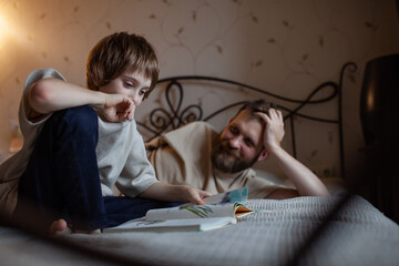 Happy boy reading book on bed while dad looking at him. Kids read with interest and pleasure. Engaging children to be readers. Parent control. Neutral colors.