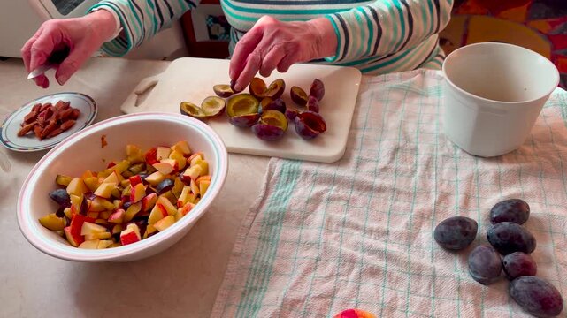 A housewife prepares a compote with plums and apples. She cuts the plums into small pieces and places the pits on a small plate. In front of her is a bowl of chopped fruit, panning shot