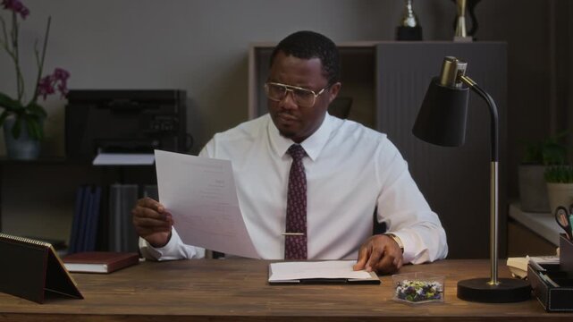 Medium portrait of male black school principal sitting at table while reviewing document and looking at camera