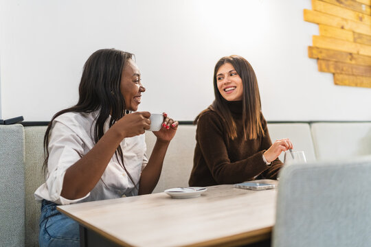 Two smiling diverse women friends enjoying coffee and having a friendly conversation at a modern cafe