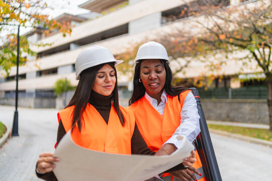 Diverse women engineers wearing hard hats and safety vests reviewing blueprints, planning a building project outdoors - Powered by Adobe