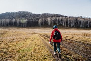 White backpacker walking into wide field toward birch grove, lone figure with red jacket and small pack, expansive view, subtle tire tracks and wilderness solitude evoke independent