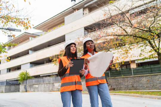 Diverse female engineers discussing new building project, holding clipboard and blueprint, standing at job site