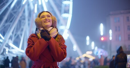 A young beautiful girl in a red coat walks in the evening at the New Year's Christmas fair against the backdrop of a Ferris wheel among lights and garlands and admires the decorations