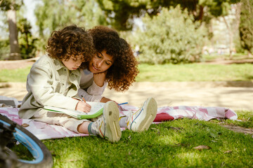 Son draws in a sketchbook with a pencil while his mother looks on and smiles as they sit on a blanket