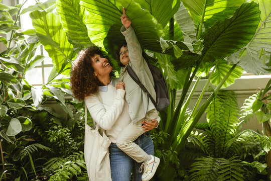 A son touches a plant leaf while his mother holds him in her arms and they smile - Powered by Adobe