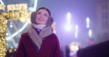 A young beautiful girl in a red coat walks in the evening at the New Year's Christmas fair against the backdrop of a Ferris wheel among lights and garlands and admires the decorations