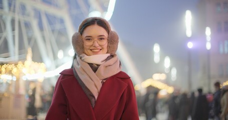A young beautiful girl in a red coat walks in the evening at the New Year's Christmas fair against the backdrop of a Ferris wheel among lights and garlands and admires the decorations
