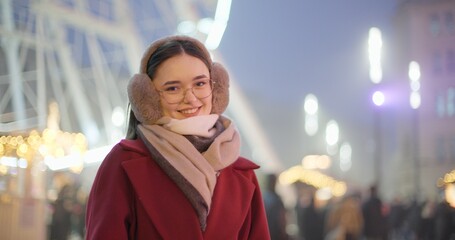 A young beautiful girl in a red coat walks in the evening at the New Year's Christmas fair against the backdrop of a Ferris wheel among lights and garlands and admires the decorations