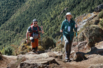 Smiling young woman hiking with male companion on rocky mountain trail in Himalayas, lush green forest slope in background.