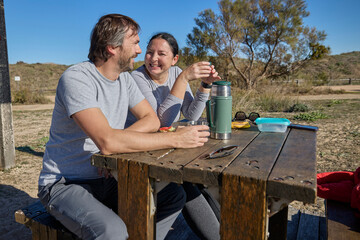 Smiling couple drinking and eating together at a rustic outdoor picnic table on a sunny day.
