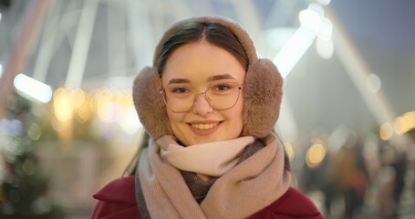 A young beautiful girl in a red coat walks in the evening at the New Year's Christmas fair against the backdrop of a Ferris wheel among lights and garlands and admires the decorations