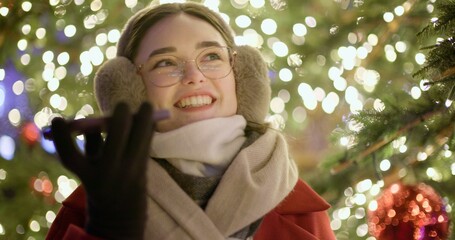 A young girl walks in the evening at the European Christmas Market, talking on the phone or reading a voice message, standing against the backdrop of a Christmas tree with garlands and illuminations.