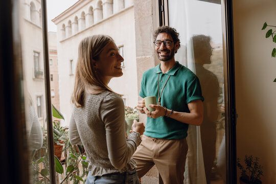 Man and woman laughing and holding cups while standing on balcony and she looking away
