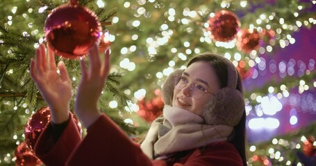 A young beautiful girl in a red coat stands near a Christmas tree with garlands and illuminations in the evening at the European Christmas Market and touches the Christmas tree decorations.