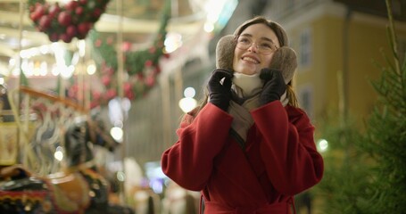 A young beautiful girl in a red coat walks in the evening at the European Christmas Market against the backdrop of a festive carousel with illuminations.