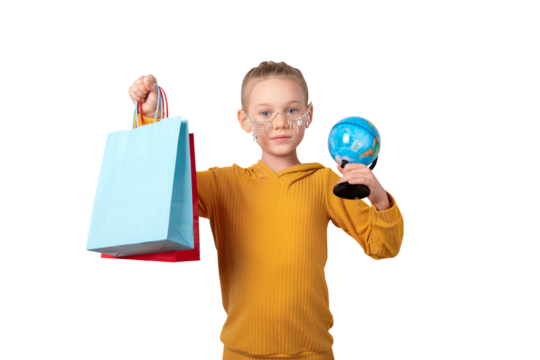 Smiling girl in yellow sweater holding colorful shopping bags and a globe, standing against a transparent background. Concept of global shopping and childhood curiosity.