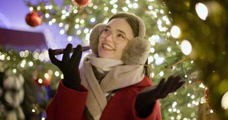 A young girl walks in the evening at the European Christmas Market, talking on the phone or reading a voice message, standing against the backdrop of a Christmas tree with garlands and illuminations.