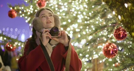 A young beautiful girl in a red coat stands near a Christmas tree with garlands and illuminations in the evening at the European Christmas Market and takes pictures with a vintage film camera.